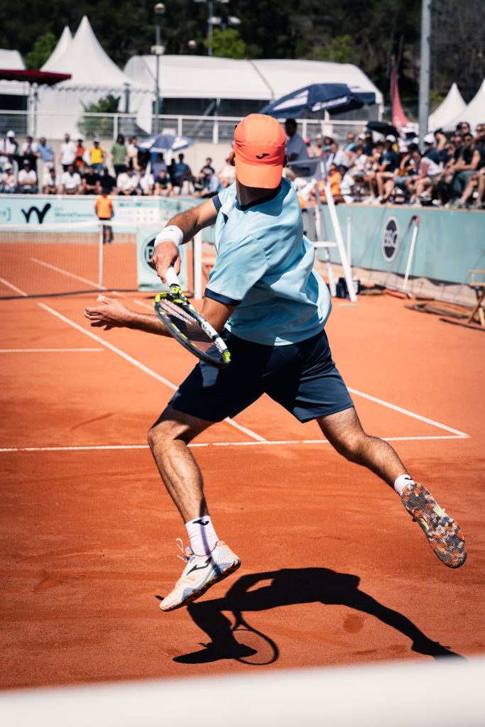 Joueur de tennis en plein coup sur terre battue lors d’un match avec public en arrière-plan.