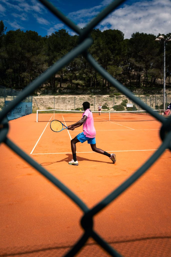 Joueur de tennis en action sur terre battue, photographié à travers un grillage, match en extérieur.