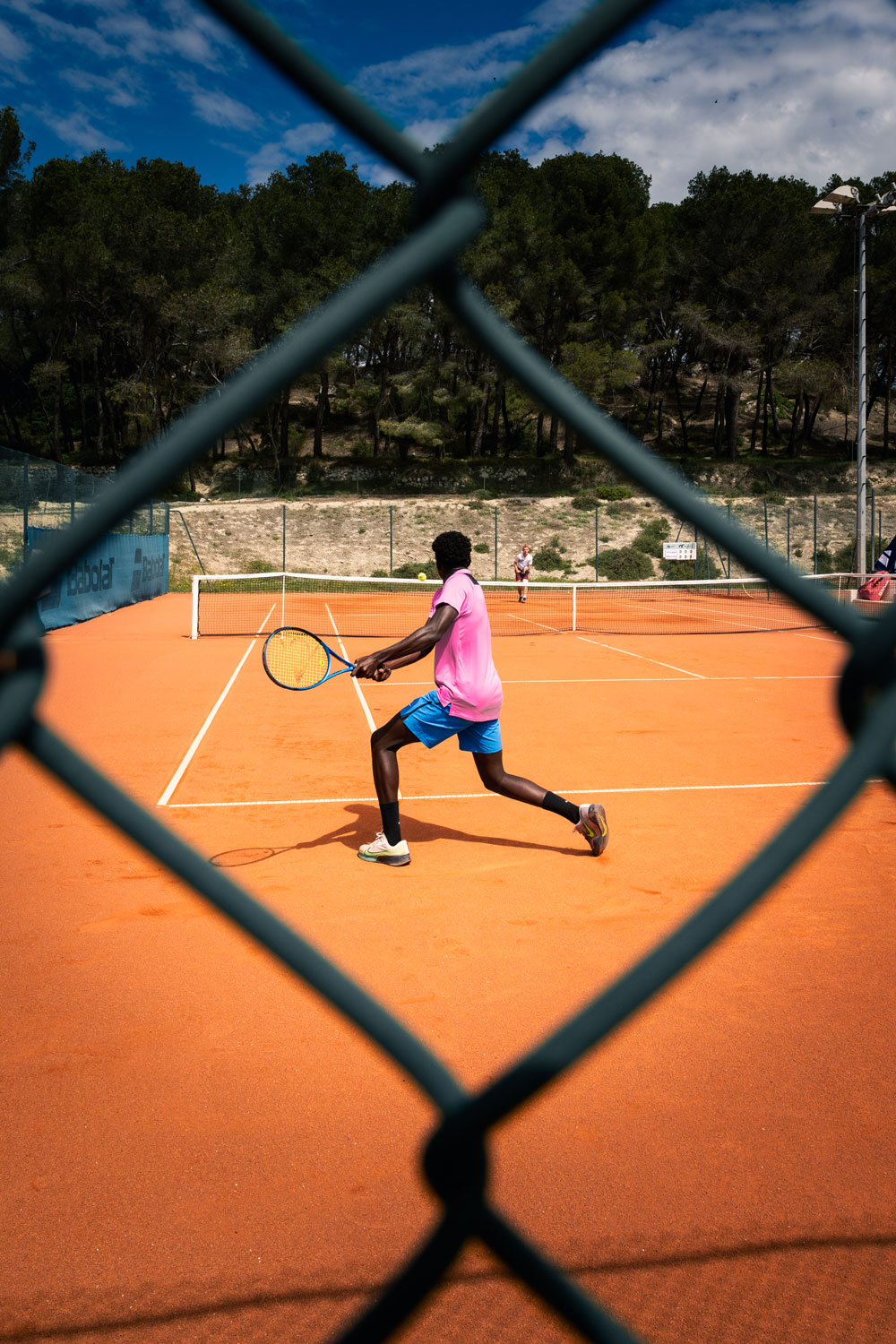 Joueur de tennis en action sur terre battue, photographié à travers un grillage, match en extérieur