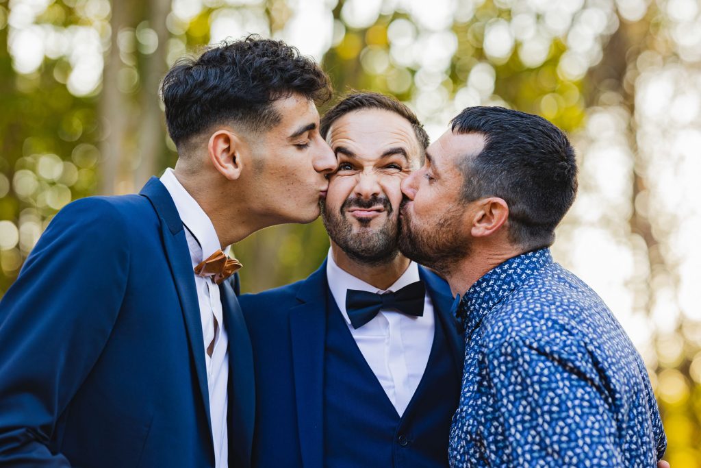 Deux invités embrassant le marié sur les joues, moment drôle et spontané lors d’un mariage en extérieur.