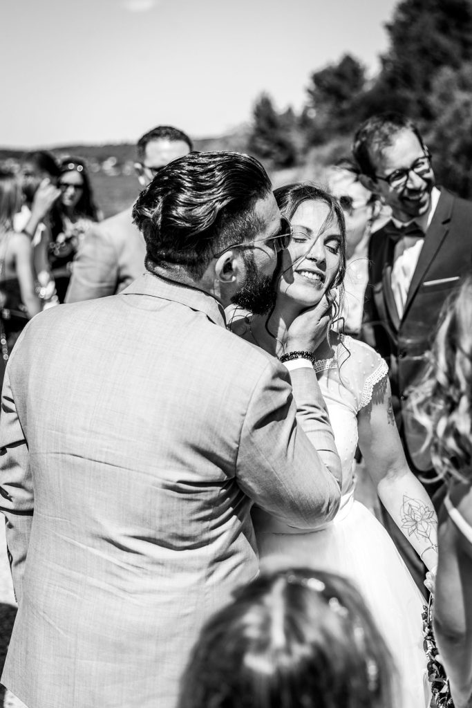 Mariés s’embrassant en extérieur entourés de leurs proches, moment tendre et spontané en noir et blanc.